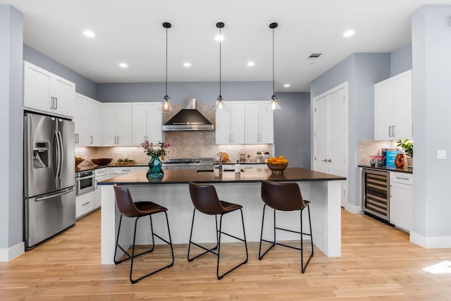 kitchen with a breakfast bar area, stainless steel appliances, wine cooler, dark countertops, and wall chimney exhaust hood