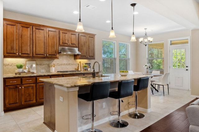 kitchen with under cabinet range hood, light stone countertops, backsplash, and brown cabinets