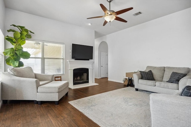 living room featuring visible vents, dark wood-type flooring, a fireplace with raised hearth, a ceiling fan, and arched walkways