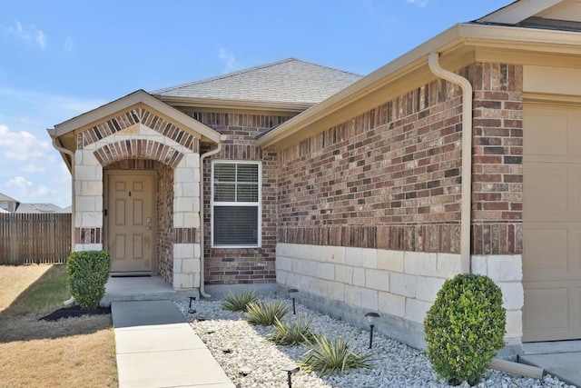 entrance to property with stone siding, fence, a shingled roof, a garage, and brick siding