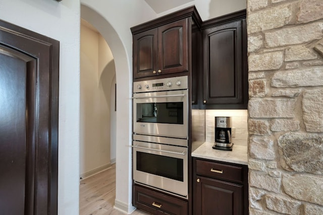 kitchen with tasteful backsplash, dark brown cabinetry, light wood-style floors, arched walkways, and stainless steel double oven