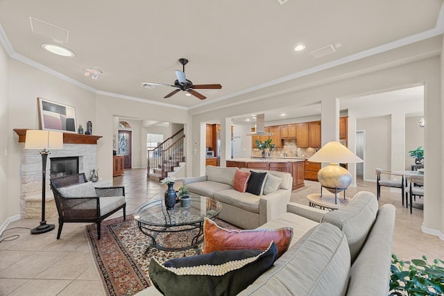 living room with light tile patterned floors, baseboards, ceiling fan, a stone fireplace, and ornamental molding