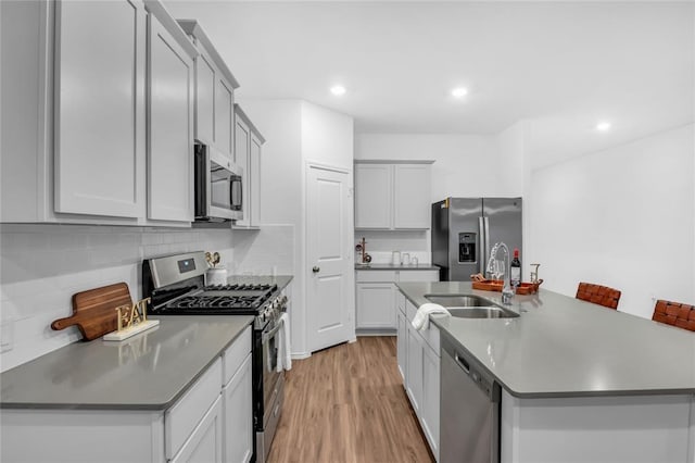 kitchen with light wood-style flooring, a kitchen island with sink, a sink, decorative backsplash, and appliances with stainless steel finishes