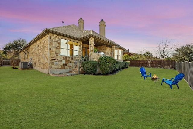 back of house at dusk featuring a yard, a fenced backyard, stone siding, and an outdoor fire pit