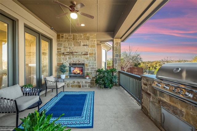 patio terrace at dusk featuring fence, exterior kitchen, ceiling fan, an outdoor stone fireplace, and a grill