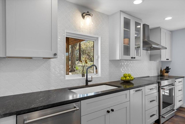 kitchen with dark stone countertops, a sink, stainless steel appliances, glass insert cabinets, and wall chimney range hood