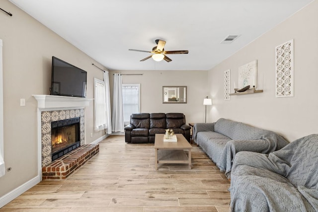 living area with visible vents, a brick fireplace, baseboards, light wood-style floors, and a ceiling fan