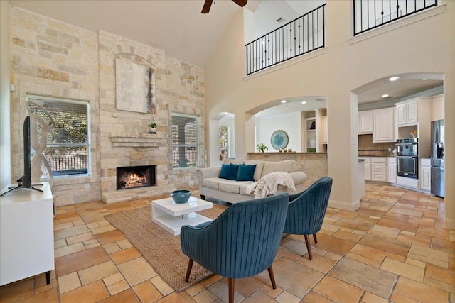 living area featuring stone tile flooring, a fireplace, a ceiling fan, and a towering ceiling