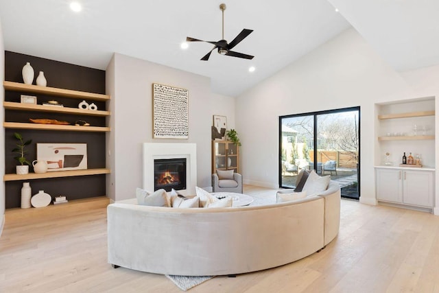 living area with ceiling fan, built in features, light wood-style floors, a glass covered fireplace, and high vaulted ceiling