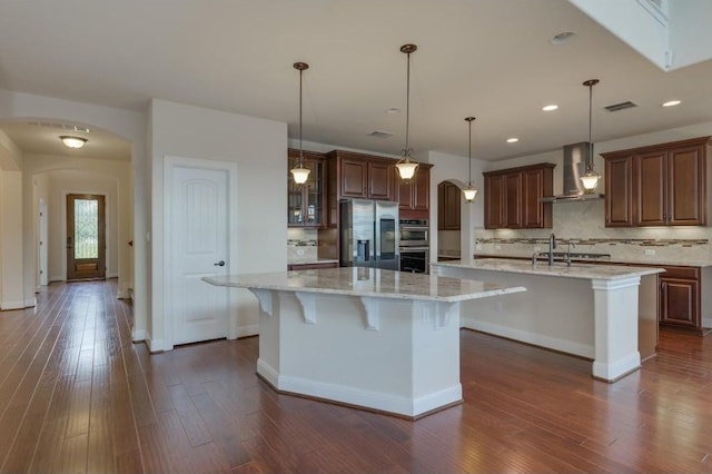 kitchen featuring a spacious island, arched walkways, stainless steel appliances, and wall chimney exhaust hood