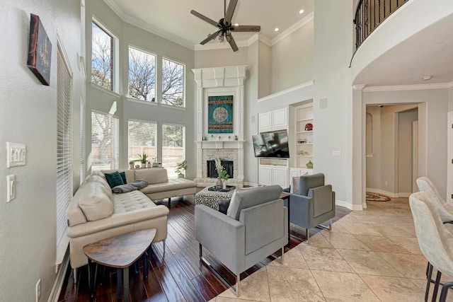 living room with built in shelves, a fireplace with raised hearth, a ceiling fan, crown molding, and baseboards