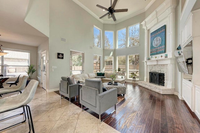 living area with wood finished floors, a stone fireplace, ceiling fan, and ornamental molding