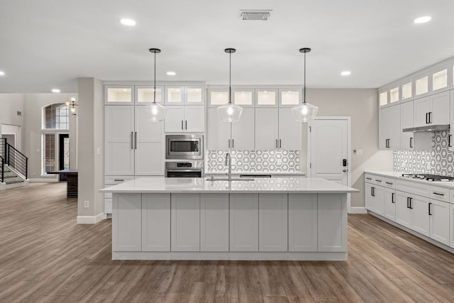 kitchen with wood finished floors, a center island with sink, a sink, light countertops, and appliances with stainless steel finishes