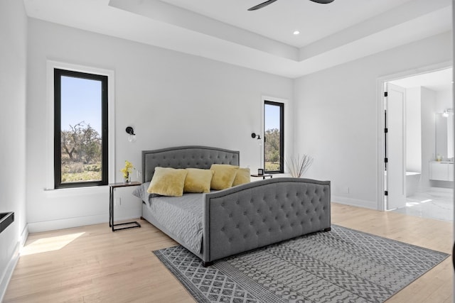 bedroom with light wood-style flooring, ensuite bathroom, a raised ceiling, and baseboards