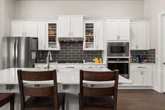 kitchen with under cabinet range hood, dark wood finished floors, decorative backsplash, stainless steel appliances, and white cabinetry