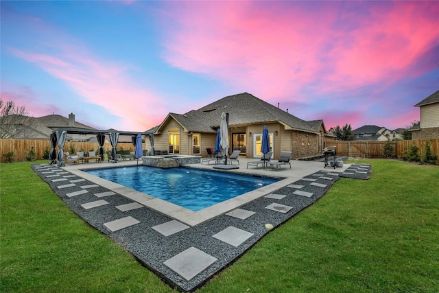 pool at dusk featuring a yard, a patio area, a pool with connected hot tub, and a fenced backyard