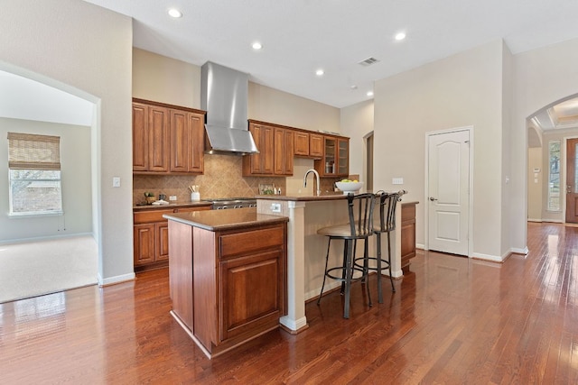 kitchen with arched walkways, brown cabinets, a kitchen island with sink, and wall chimney range hood