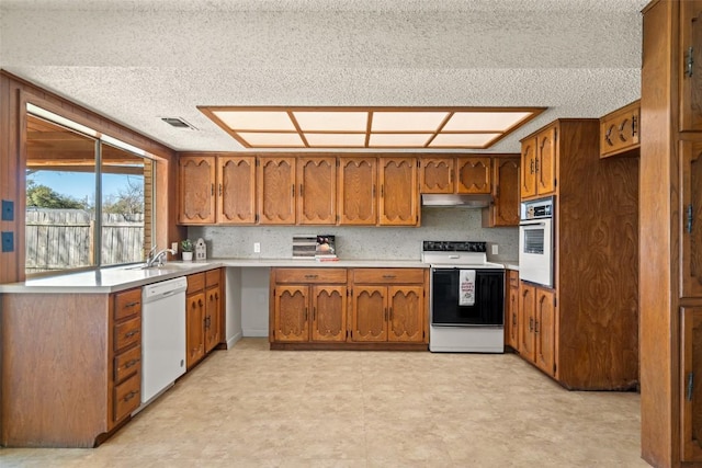 kitchen with tasteful backsplash, under cabinet range hood, light countertops, brown cabinets, and white appliances