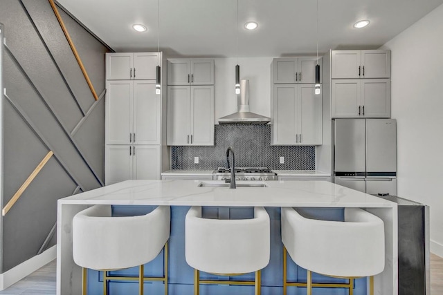 kitchen with light stone countertops, an island with sink, freestanding refrigerator, wall chimney exhaust hood, and tasteful backsplash