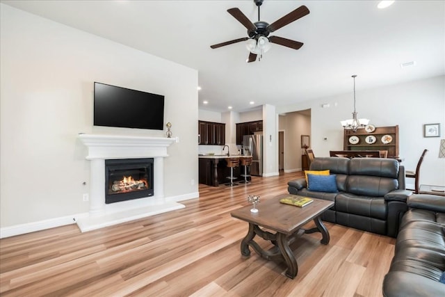 living room featuring light wood-style flooring, ceiling fan with notable chandelier, a glass covered fireplace, recessed lighting, and baseboards