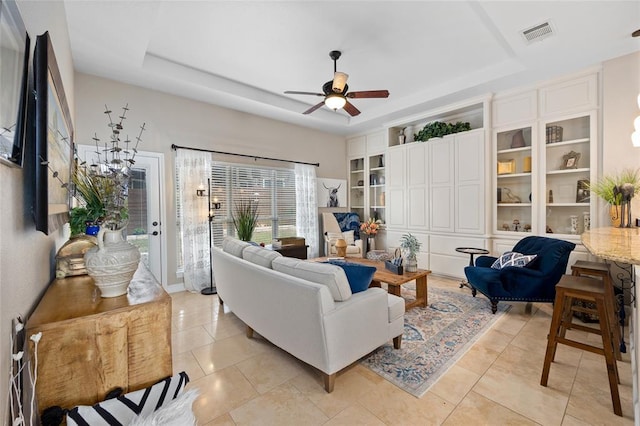living room featuring a raised ceiling, visible vents, light tile patterned flooring, built in shelves, and ceiling fan