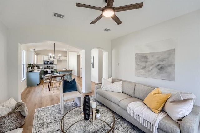 living area featuring ceiling fan with notable chandelier, light wood-style floors, visible vents, and arched walkways