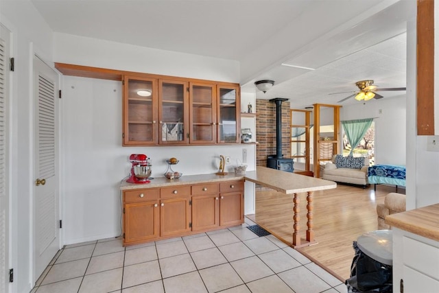 kitchen with brown cabinetry, a wood stove, ceiling fan, light countertops, and glass insert cabinets