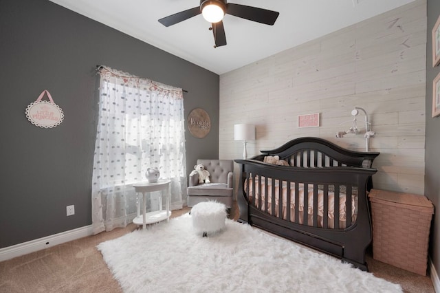 carpeted bedroom featuring baseboards, a nursery area, ceiling fan, and wooden walls