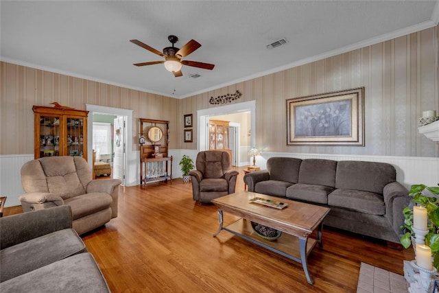 living room featuring a wainscoted wall, visible vents, light wood-style flooring, crown molding, and ceiling fan