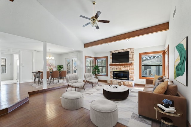 living area with visible vents, beamed ceiling, a stone fireplace, ceiling fan with notable chandelier, and wood finished floors