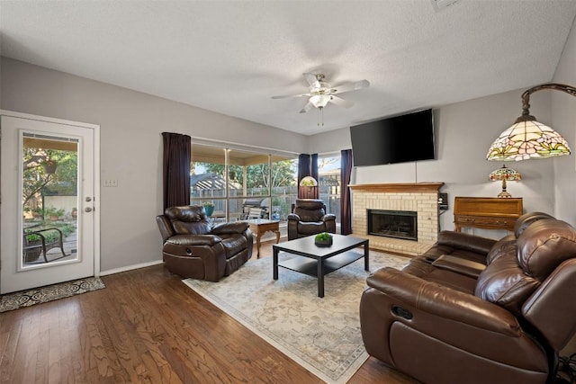 living room featuring a brick fireplace, dark wood-type flooring, baseboards, a textured ceiling, and a ceiling fan