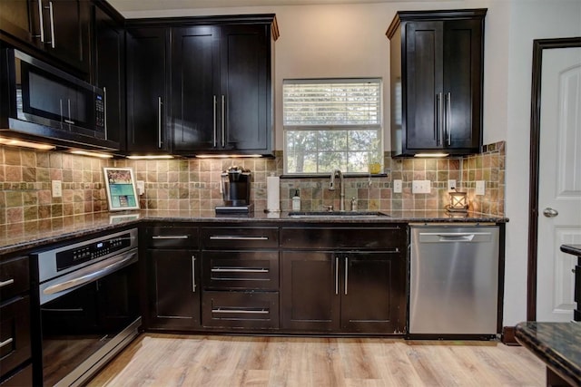 kitchen with a sink, dark stone counters, appliances with stainless steel finishes, and light wood finished floors