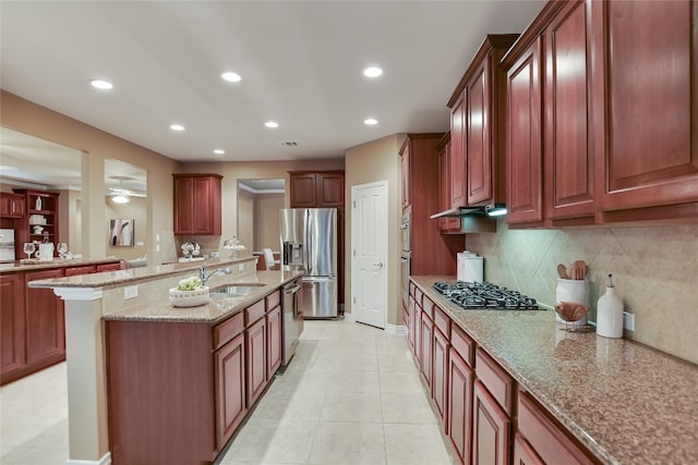 kitchen featuring a center island with sink, a sink, light stone counters, stainless steel appliances, and decorative backsplash