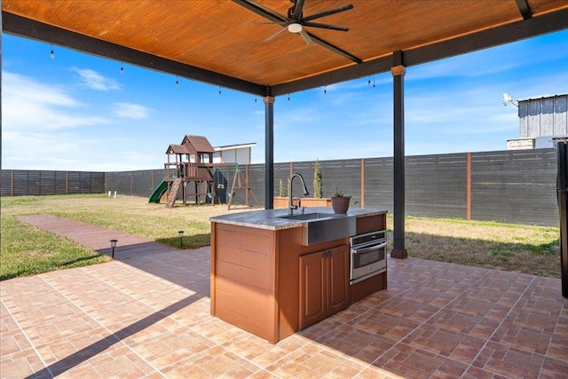 view of patio with a playground, exterior kitchen, a fenced backyard, a ceiling fan, and a sink