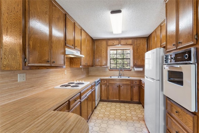 kitchen with white appliances, brown cabinetry, a sink, light countertops, and under cabinet range hood