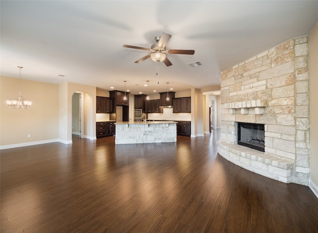 unfurnished living room featuring visible vents, a stone fireplace, ceiling fan with notable chandelier, arched walkways, and dark wood-style floors