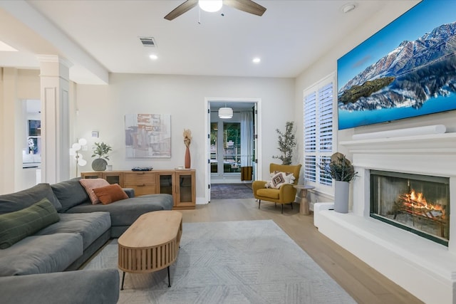 living room with wood finished floors, a ceiling fan, visible vents, ornate columns, and a lit fireplace