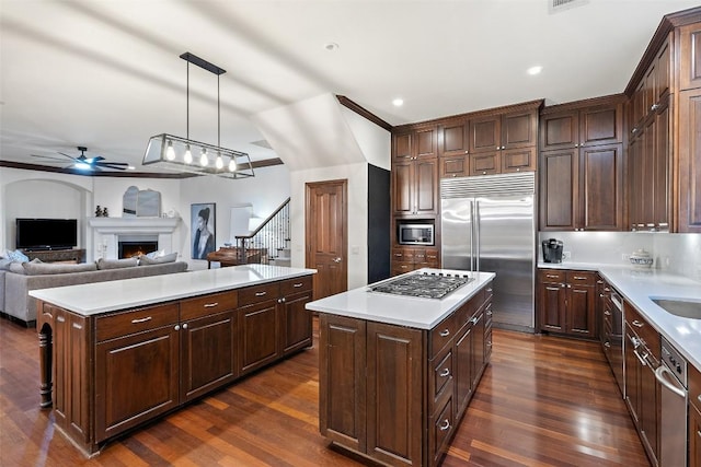 kitchen with a ceiling fan, a warm lit fireplace, dark wood-type flooring, built in appliances, and a center island