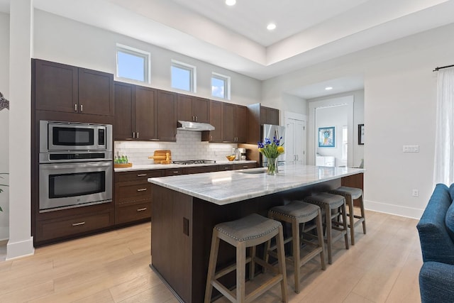 kitchen with tasteful backsplash, dark brown cabinets, under cabinet range hood, a kitchen breakfast bar, and stainless steel appliances