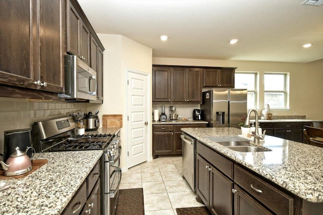 kitchen with an island with sink, a sink, dark brown cabinets, appliances with stainless steel finishes, and tasteful backsplash