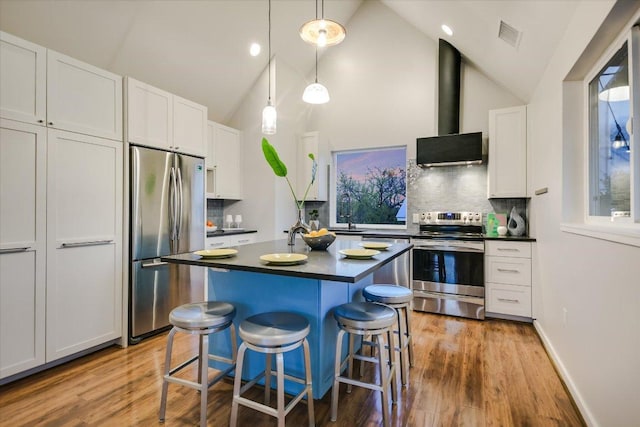 kitchen with dark countertops, light wood finished floors, a kitchen breakfast bar, stainless steel appliances, and wall chimney exhaust hood