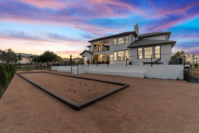 back of house at dusk featuring stucco siding, a balcony, fence, and a chimney