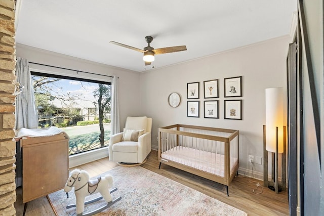 bedroom with a nursery area, a ceiling fan, baseboards, and wood finished floors