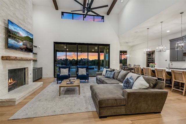 living area with beam ceiling, light wood-style floors, a fireplace, and ceiling fan with notable chandelier