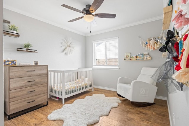 bedroom with wood finished floors, baseboards, ceiling fan, a crib, and ornamental molding