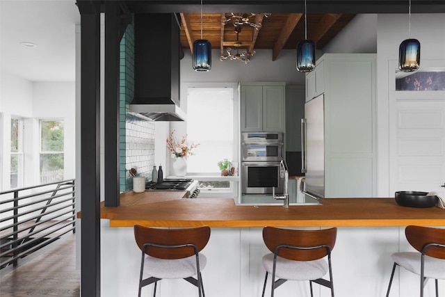 kitchen featuring beam ceiling, decorative light fixtures, a kitchen breakfast bar, double oven, and wall chimney range hood