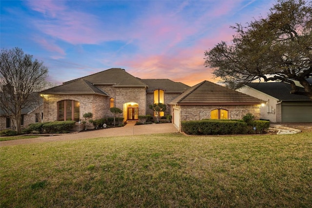 french country home featuring driveway, an attached garage, stone siding, a tile roof, and a lawn