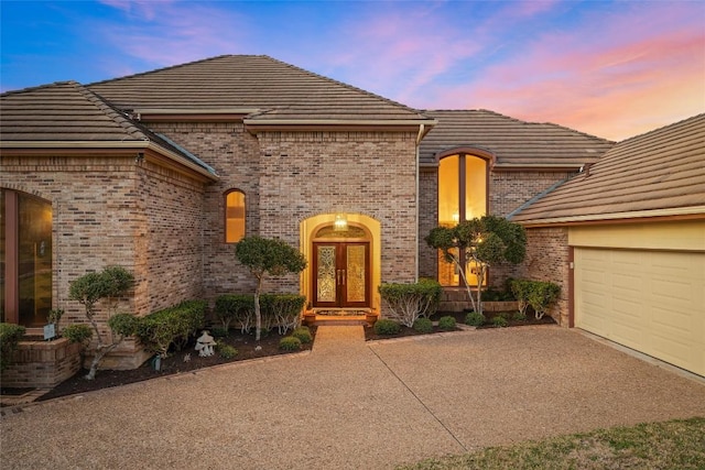 french country style house featuring brick siding, a tiled roof, an attached garage, and driveway