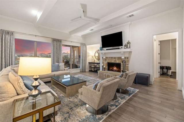 living room with beamed ceiling, wood finished floors, a stone fireplace, crown molding, and ceiling fan