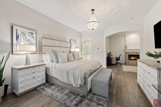 bedroom with arched walkways, a stone fireplace, baseboards, and wood finished floors